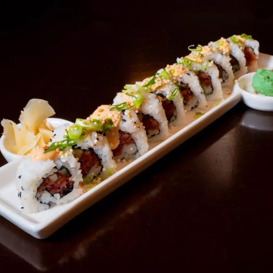 A long white plate of sushi rolls with tuna, sauce, green onions, and sesame seeds.