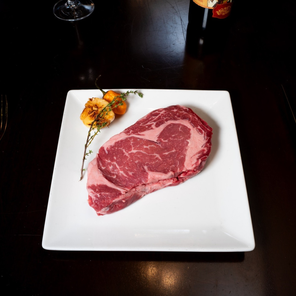 Uncooked ribeye steak on a white plate with roasted garlic, peppers, and thyme sprigs.