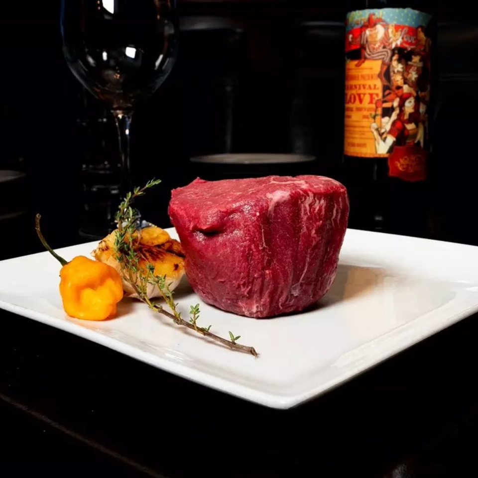 Raw steak filet on a white plate with a yellow pepper, herbs, and a wine bottle.