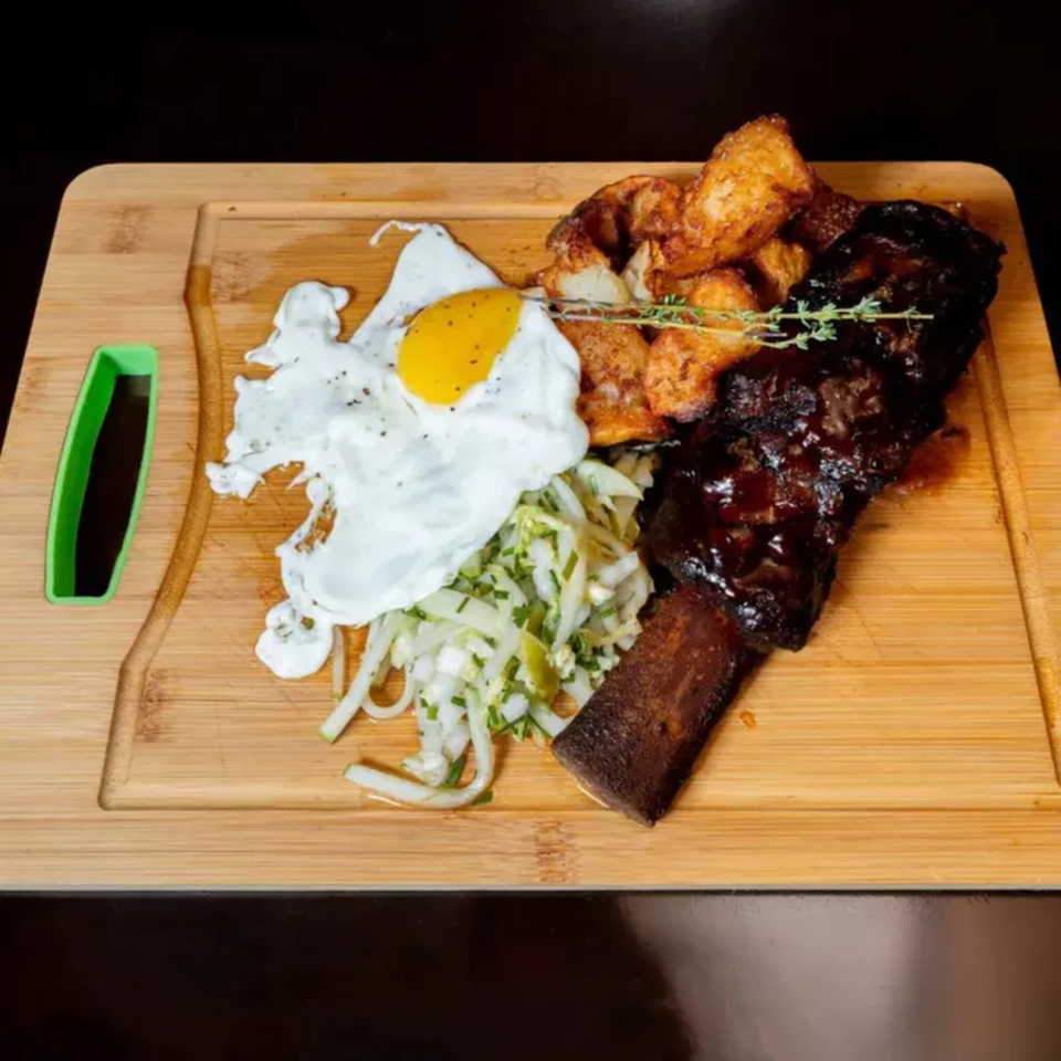 A Polo Steak and Sea short rib with fried egg, potatoes, and coleslaw on a wooden board.