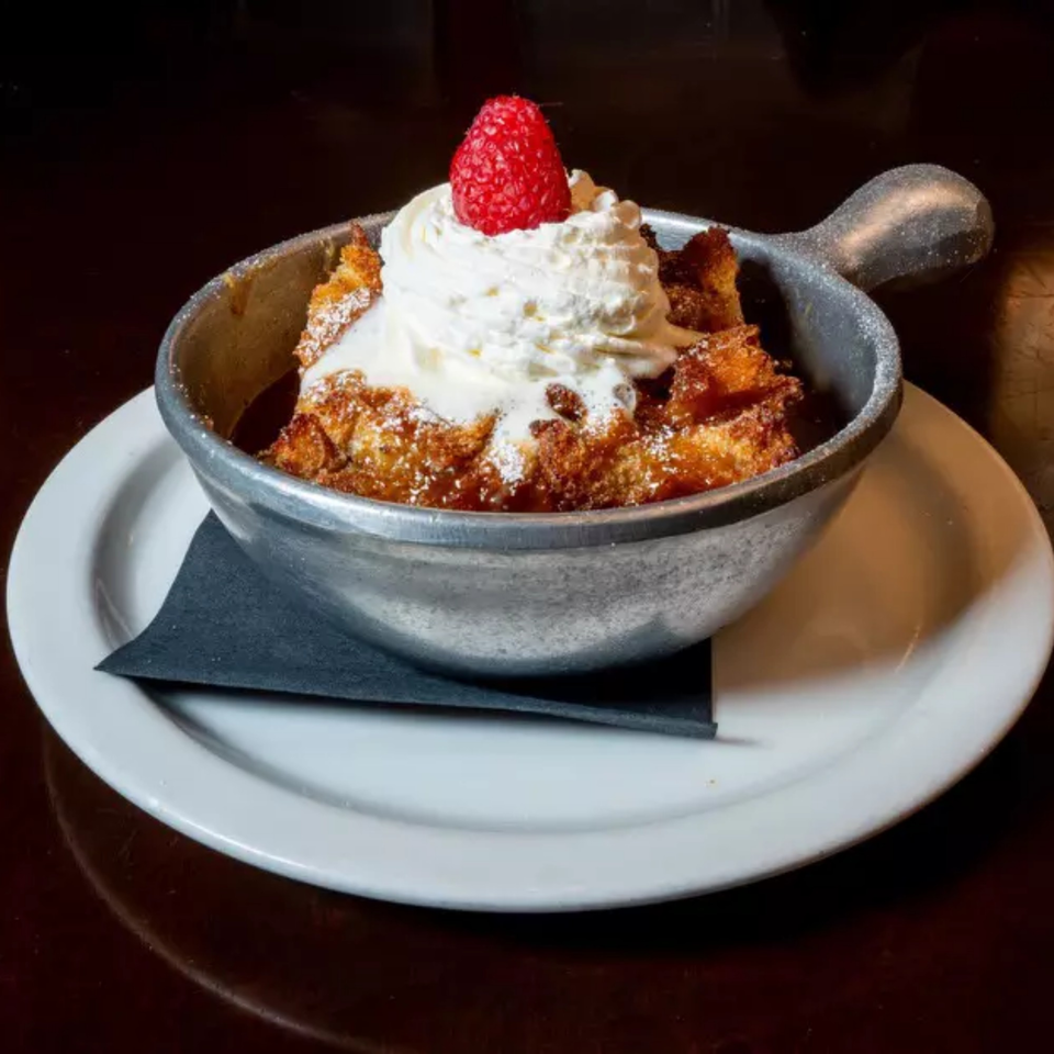 Bread pudding in a metal skillet, topped with whipped cream and a fresh raspberry.